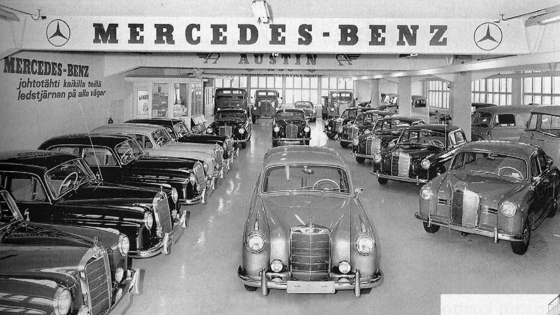 Mustavalkoinen kuva autoliikkeestä 1950-luvulla. Mercedes-Benz autot rivissä vasemmalla ja keskellä yksi auto kuvattuna edestä päin.